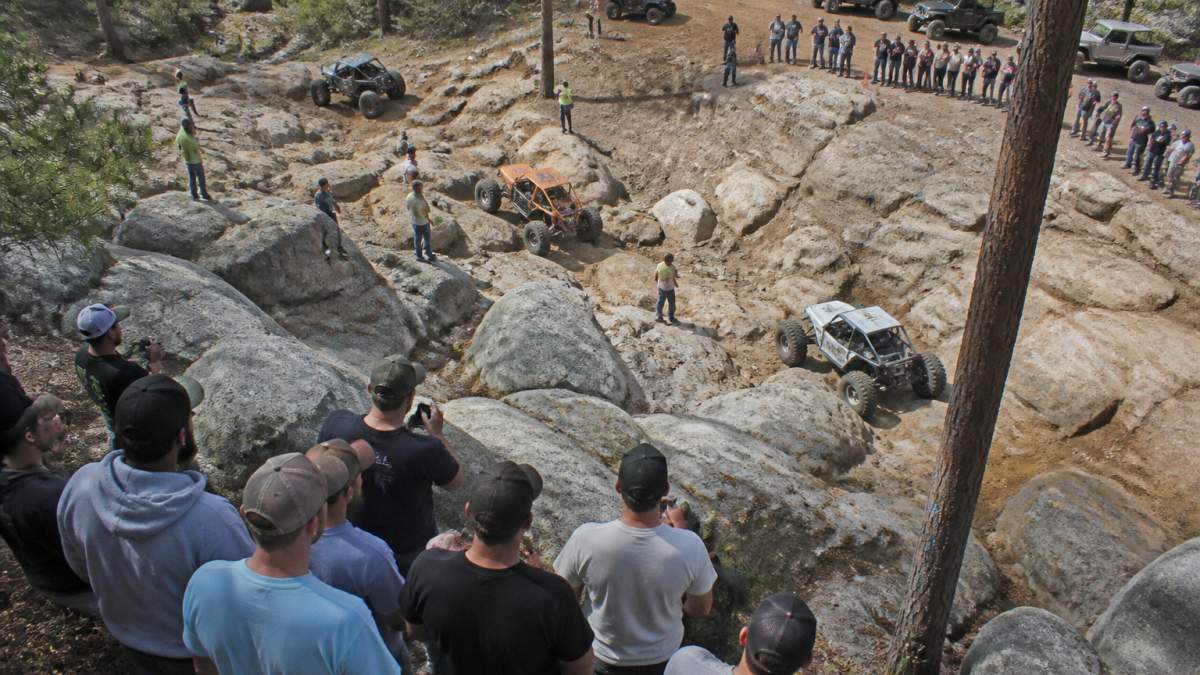 Overhead view of a rock crawling course with multiple vehicles navigating rocky obstacles, spectators watching from designated viewing areas, and the course layout visible with steep rocky terrain and natural boulder formations