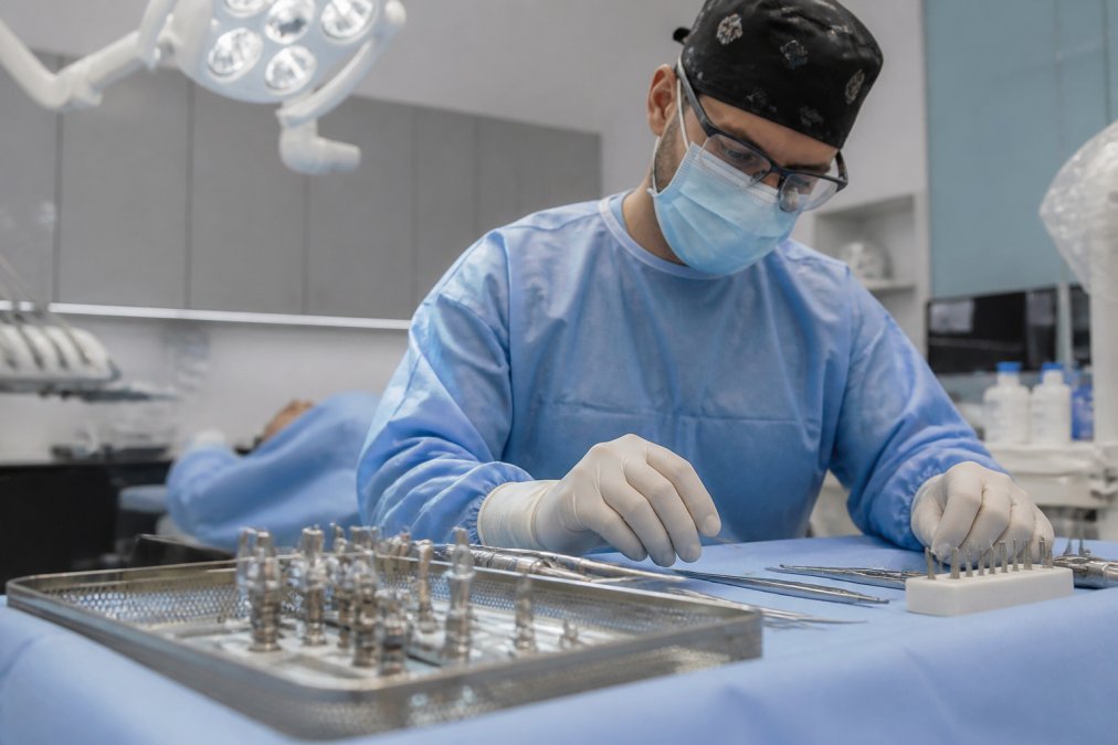 Dental surgeon in surgical mask and protective eyewear preparing sterilized titanium implant instruments on a surgical tray in a modern operating room with overhead surgical lighting