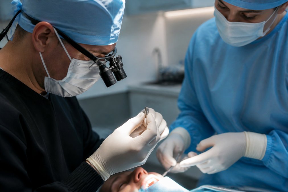 Close-up of a dentist's gloved hands holding a small titanium implant post under bright surgical lighting in a modern treatment room