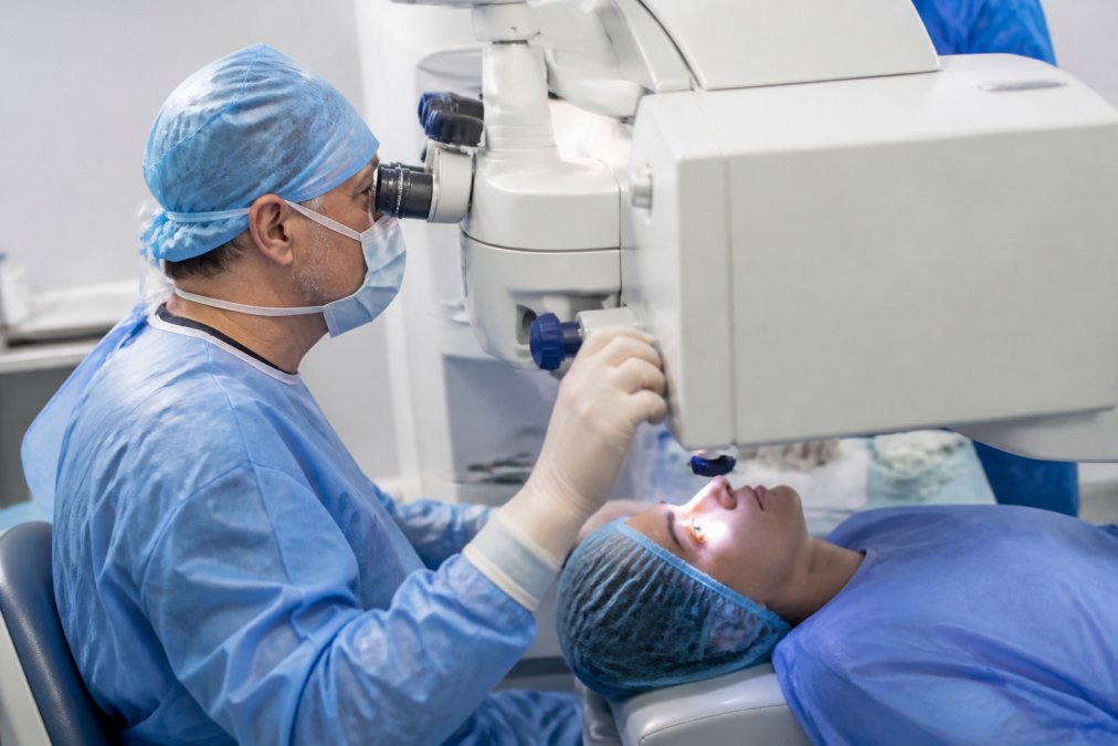 Patient positioned in LASIK procedure chair with eyelid holder in place, surgeon adjusting laser equipment, clinical environment with soft overhead lighting and monitor displays