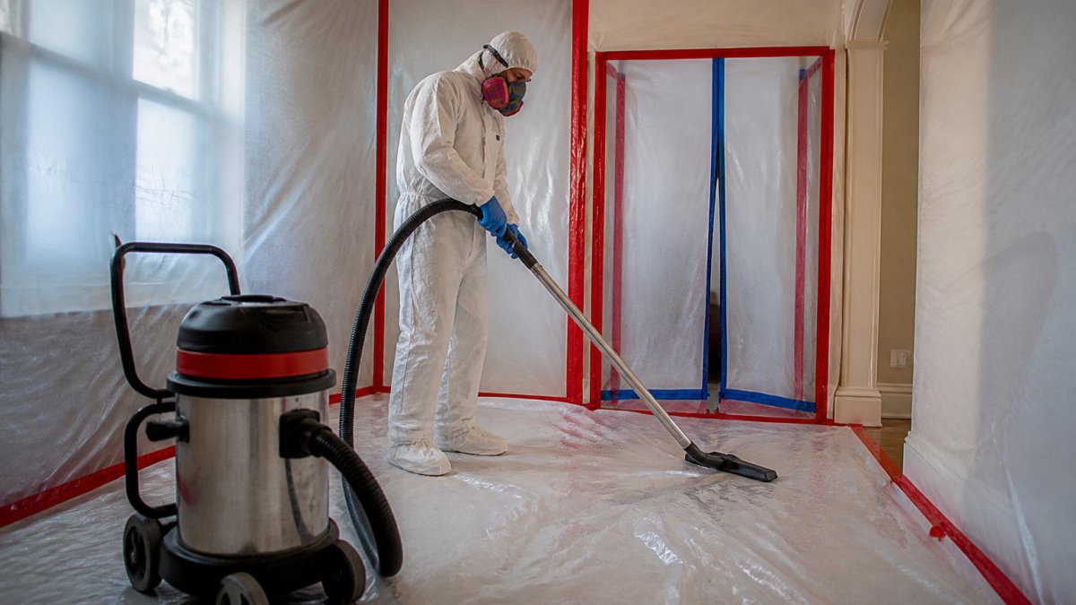 Professional demonstrating lead abatement containment setup with protective plastic sheeting on walls and floors, HEPA vacuum equipment, and worker in full protective gear with respirator in modern home interior