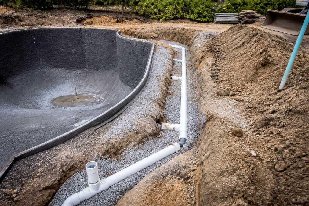 Close-up view of drainage pipes and grading work around an excavated pool area with water management infrastructure being installed and soil layers visible