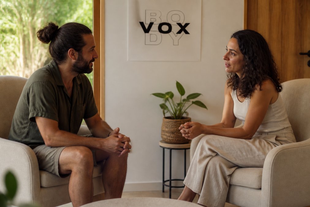 Two people sitting face-to-face in a softly lit room with comfortable seating, engaged in attentive conversation with calm, open body language and relaxed posture