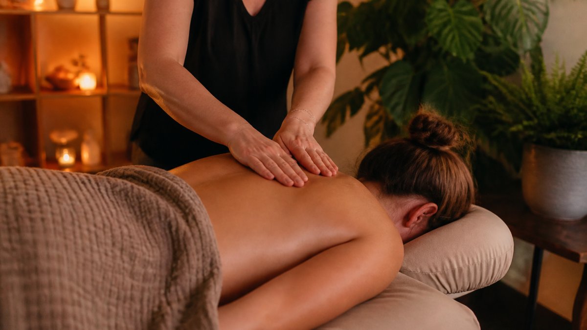Person lying face-down on a massage table in a softly lit room with warm ambient lighting, plants visible in background, therapist's hands visible working on upper back