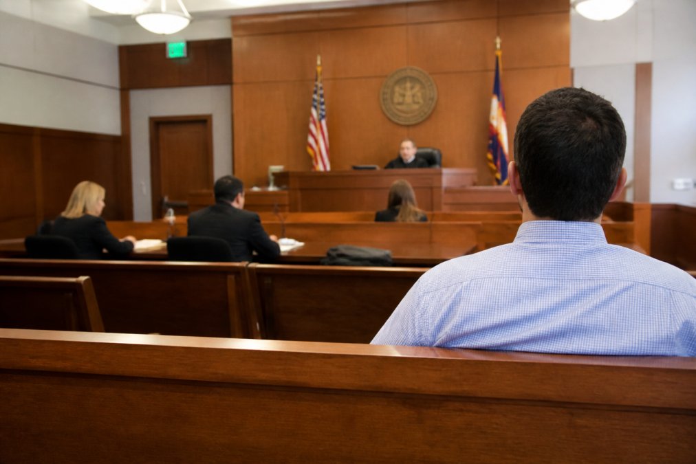 Individual sitting in a courtroom gallery observing proceedings, showing the typical courtroom environment with the judge's elevated bench, attorney tables, and organized seating arrangement