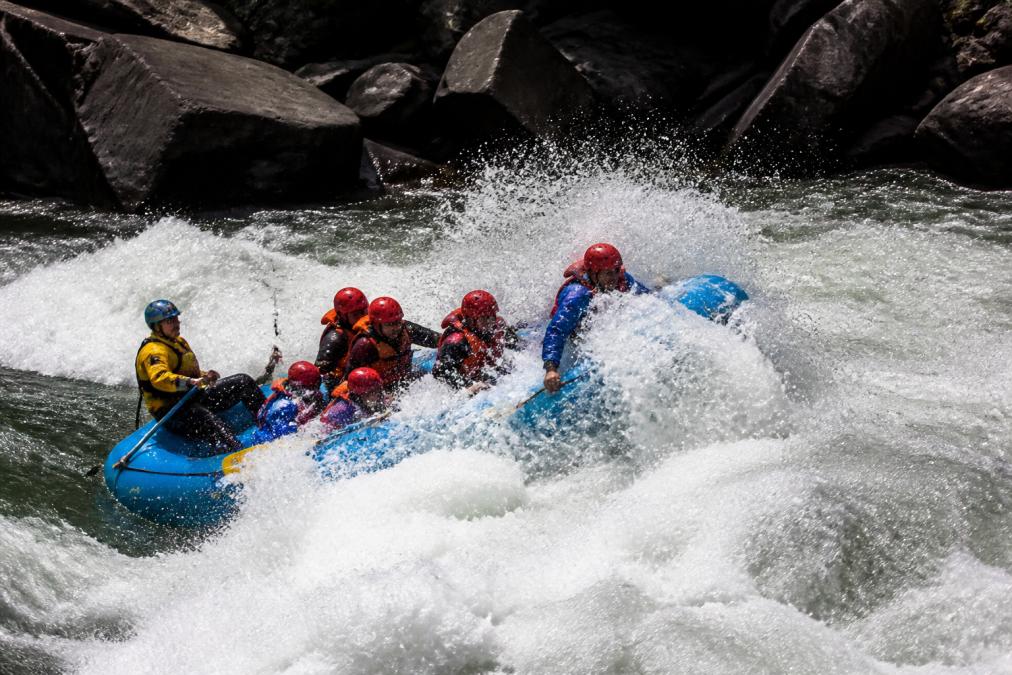 A commercial raft navigating through massive white water and towering waves during an intense Class V rapid, with paddlers gripping safety ropes and spray flying across the boat, showing the violent and chaotic nature of the whitewater