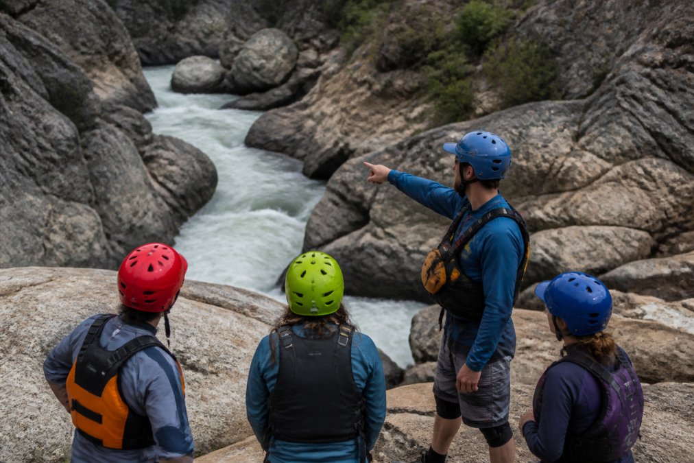 A professional whitewater guide and paddlers standing on shore examining the rapid from above, studying the line and identifying hazards before running the Class V section, demonstrating the critical importance of scouting