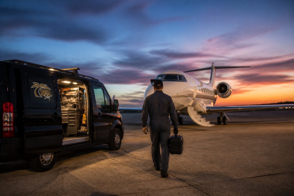 A mobile aircraft maintenance van parked beside a private jet on a South Florida tarmac at dusk, with a technician in coveralls walking toward the aircraft carrying a tool case