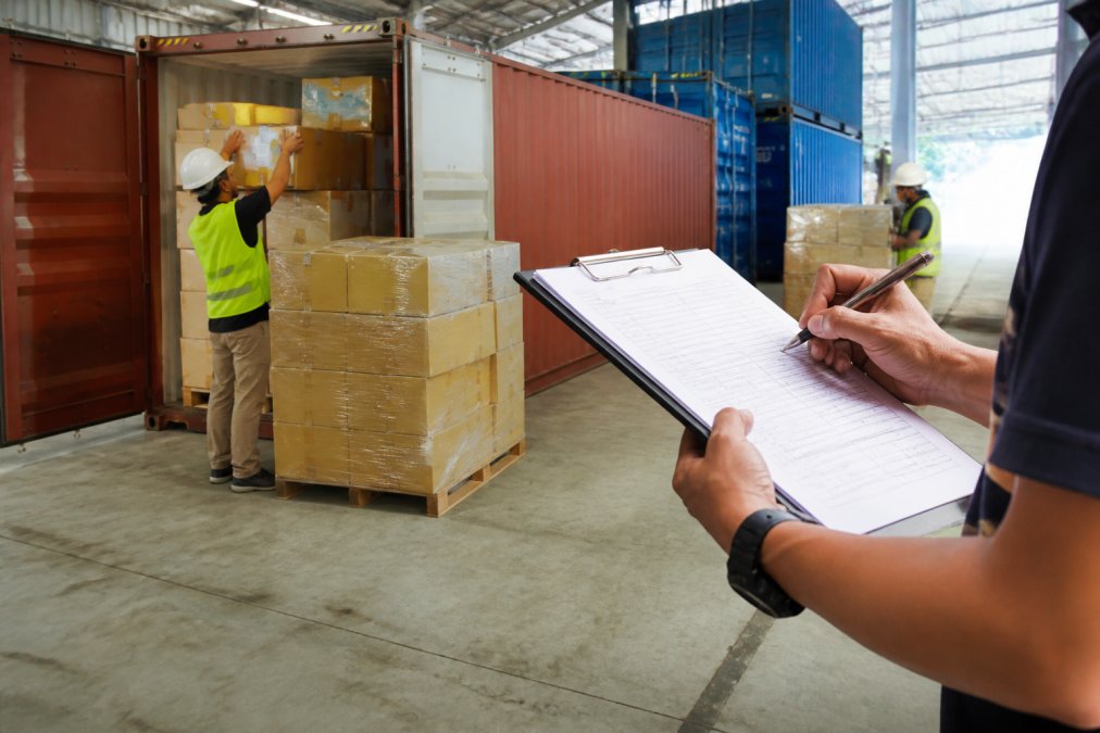 Logistics warehouse with workers managing containerized cargo stacked in rows, freight documentation clipboard in foreground, natural warehouse lighting