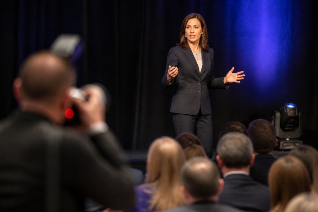 Professional photographer capturing a keynote speaker at a corporate conference with proper lighting setup, clear focus on the speaker's expression and gesture, demonstrating technical control in challenging venue lighting conditions