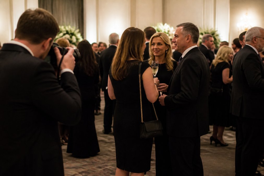 Professional event photographer positioned at edge of corporate gala, discreetly capturing candid moments between attendees without being intrusive, evening lighting with soft background blur