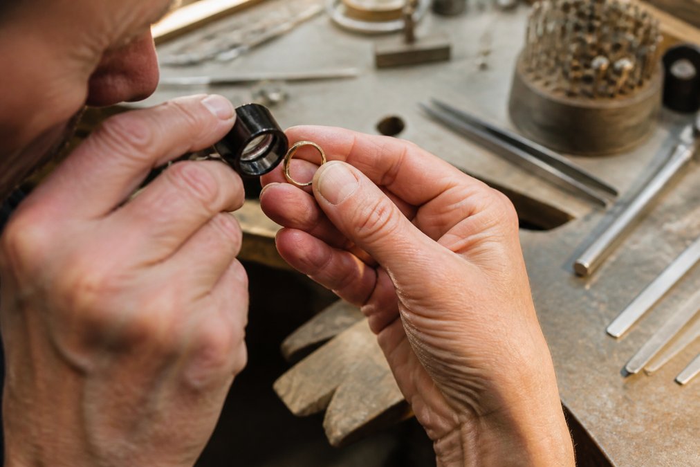 Close-up of a jeweler's hands holding a jeweler's loupe to inspect the inside of a vintage gold ring, with a soft-focus background of a workbench