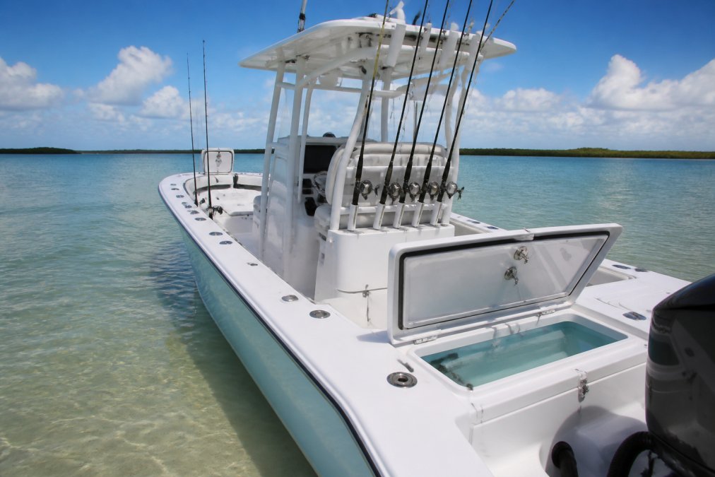 Wide-angle view of center console fishing boat at anchor in shallow coastal waters with multiple rod holders visible along the gunwale, live well hatch open in foreground, and calm offshore waters stretching to horizon
