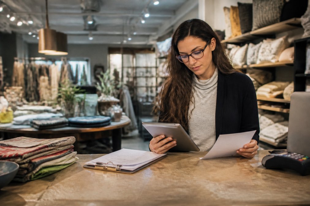 A small business owner reviewing insurance documents on a tablet inside their retail store, with shelves of inventory and display fixtures visible in the background under bright overhead lighting