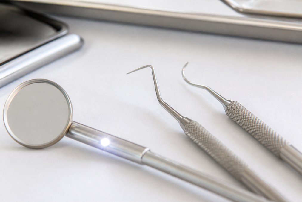 Close-up of stainless steel dental instruments including mouth mirror with LED light, explorer probe, and periodontal scaler arranged on a clean white surface with soft clinical lighting