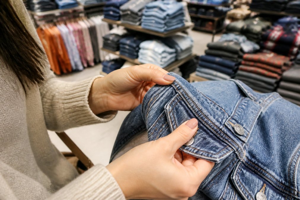 Close-up of a woman's hands inspecting the stitching and fabric texture of a folded denim jacket in a brightly lit discount retail store