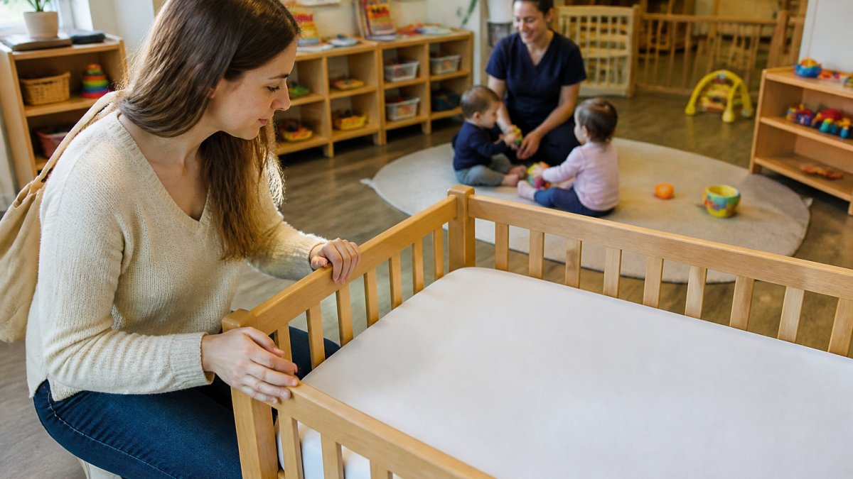Parent inspecting a crib at an infant daycare while a caregiver supervises babies playing on a mat nearby.