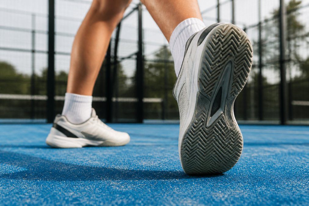 Close-up of padel shoe sole showing detailed herringbone tread pattern in contact with padel court surface, with player's foot positioned mid-lateral movement demonstrating the shoe's grip and support during directional change