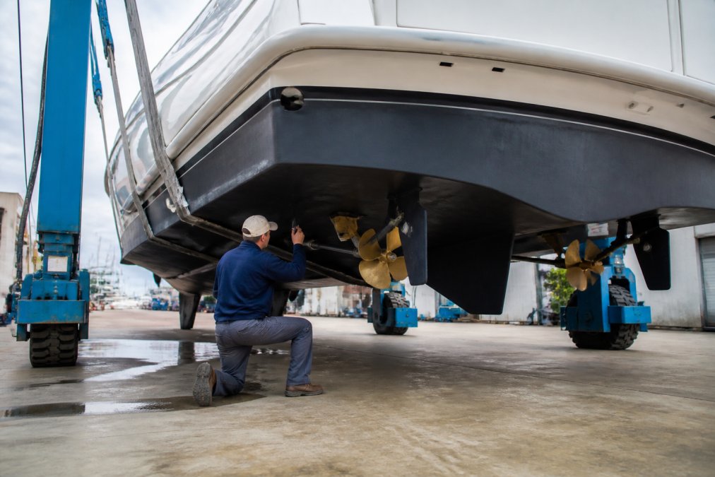 A marine surveyor in work clothes inspecting the flared hull and running gear of a large sportfish yacht hauled out on a boatyard lift, with the yacht's transom and propeller shafts visible in daylight