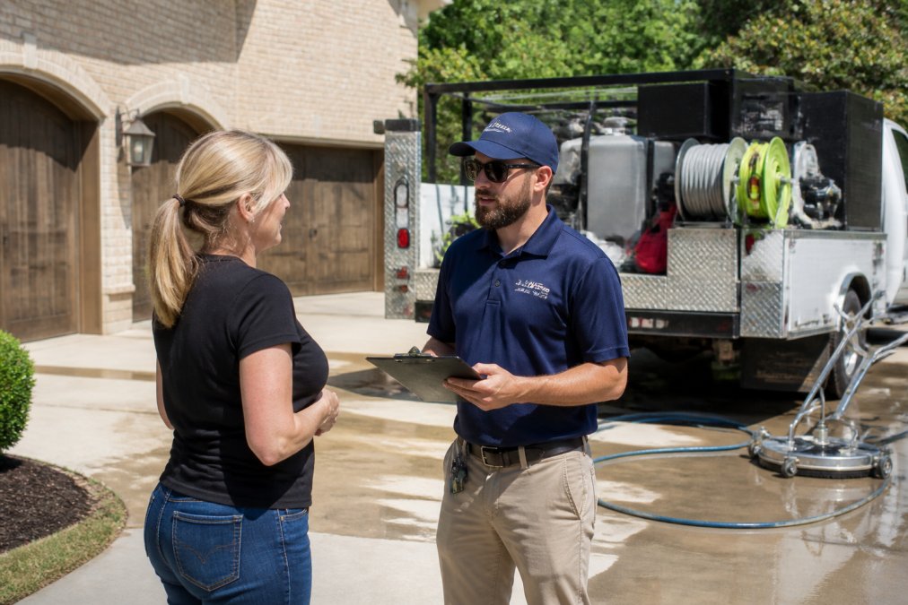A homeowner standing in a driveway talking with a pressure washing technician who is holding a clipboard, with a pressure washing truck and equipment visible in the background on a sunny day