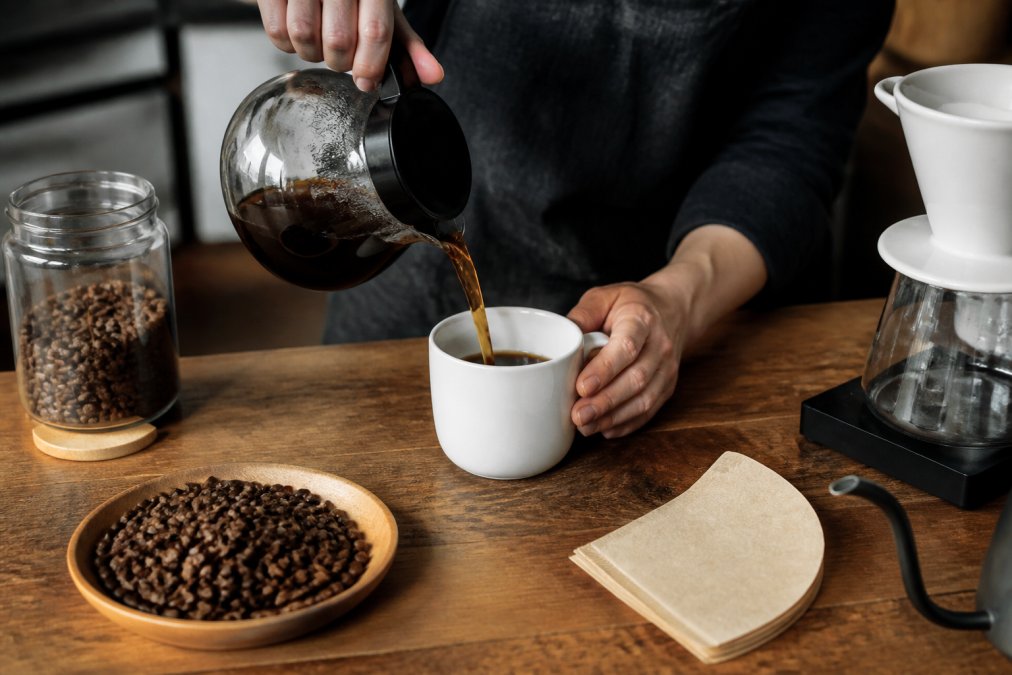 Freshly brewed black coffee being poured into a white ceramic mug on a wooden counter with coffee beans.