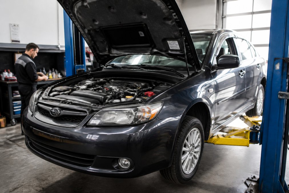 Well-maintained Japanese sedan on lift in professional repair shop with clean engine bay, technician in background performing inspection, organized tools and equipment visible in modern facility