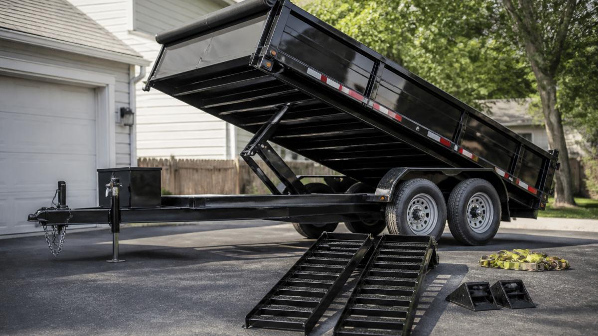 12-yard dump trailer parked on residential driveway showing open hydraulic bin, loading ramps positioned at side, tie-down straps coiled nearby, and wheel chocks on ground in daylight