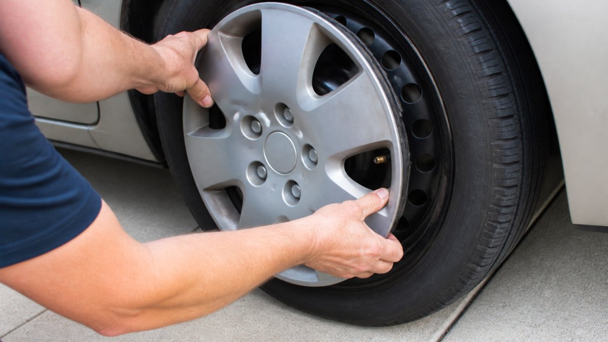 Close-up of two hands pressing a plastic wheel cover onto a steel car wheel, valve stem aligned with the cover's cutout, driveway setting, natural daylight
