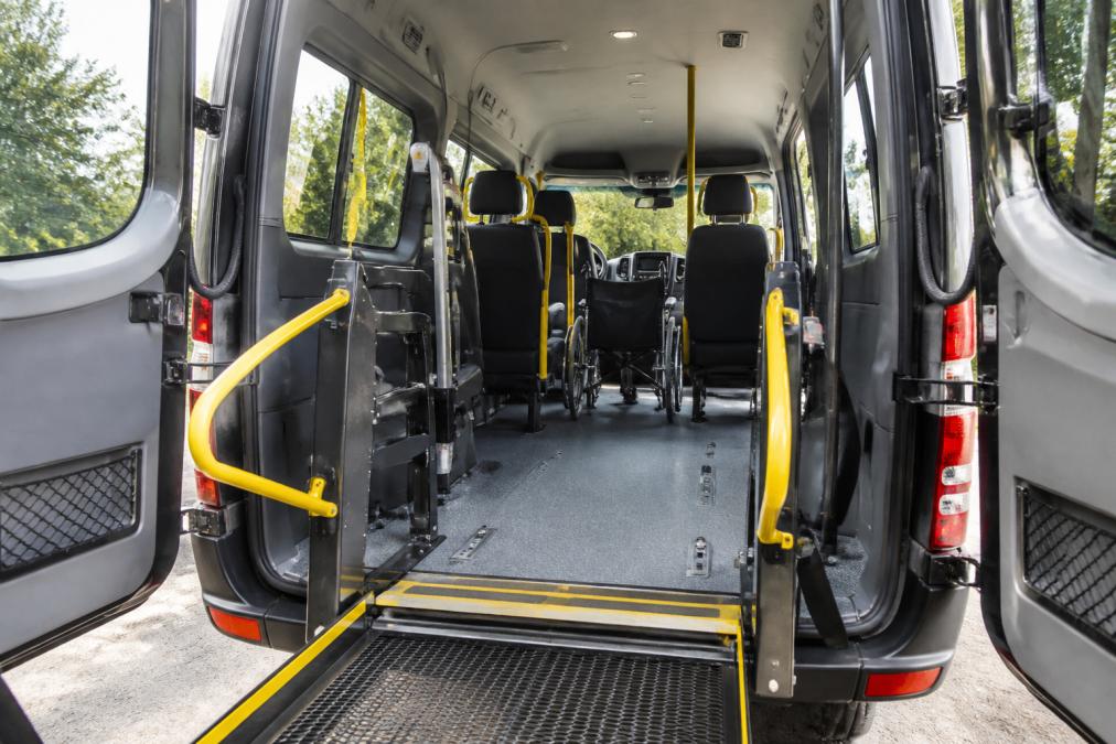Interior view of a modern wheelchair accessible minibus with deployed wheelchair lift, accessible seating arrangements, and safety railings visible in natural daylight