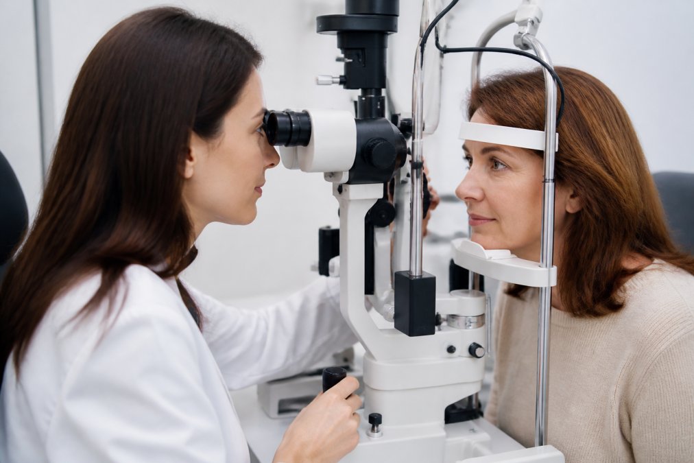 An eye care professional in a bright exam room gently examining a middle-aged patient's eye with a slit lamp, both wearing a calm and reassuring expression