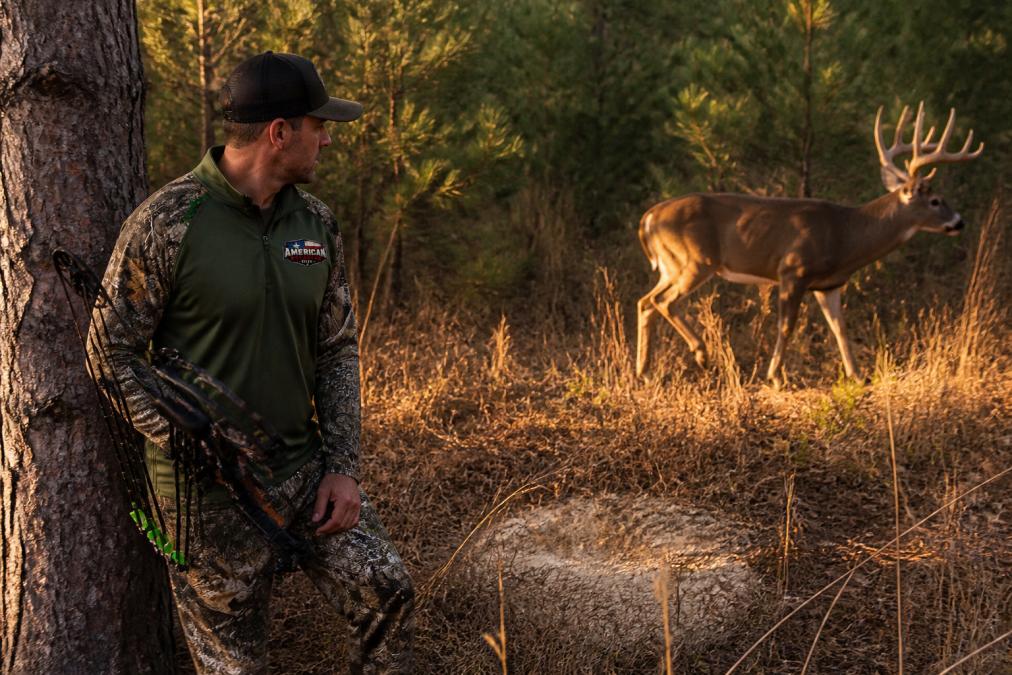 A large whitetail buck with a full antler rack moving along a forest edge at low golden light, a fresh scrape visible on the ground nearby, pine trees in the background