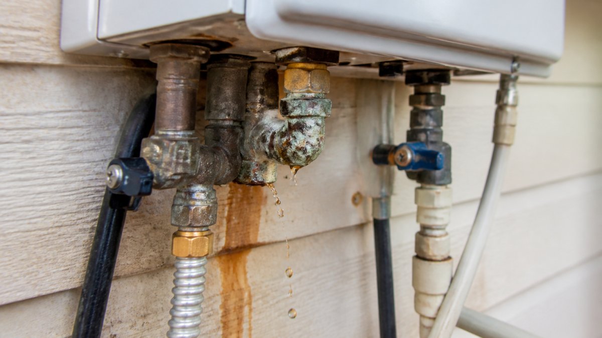 Close-up of water dripping from a corroded pipe connection on a tankless water heater unit mounted on a wall, showing rust stains and mineral deposits