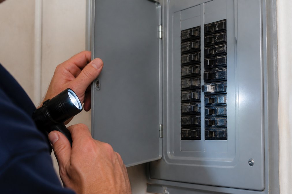 Close-up of a homeowner's hands opening an electrical panel door, flashlight in one hand, inspecting a row of circuit breakers with slight scorch marks visible on one switch