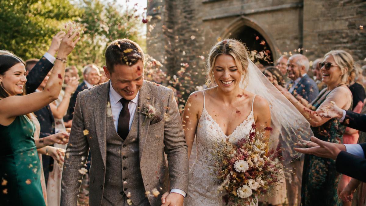 A newlywed couple walking through a shower of dried flower petal confetti thrown by smiling guests outside a stone church, golden afternoon light, petals suspended mid-air, candid documentary style