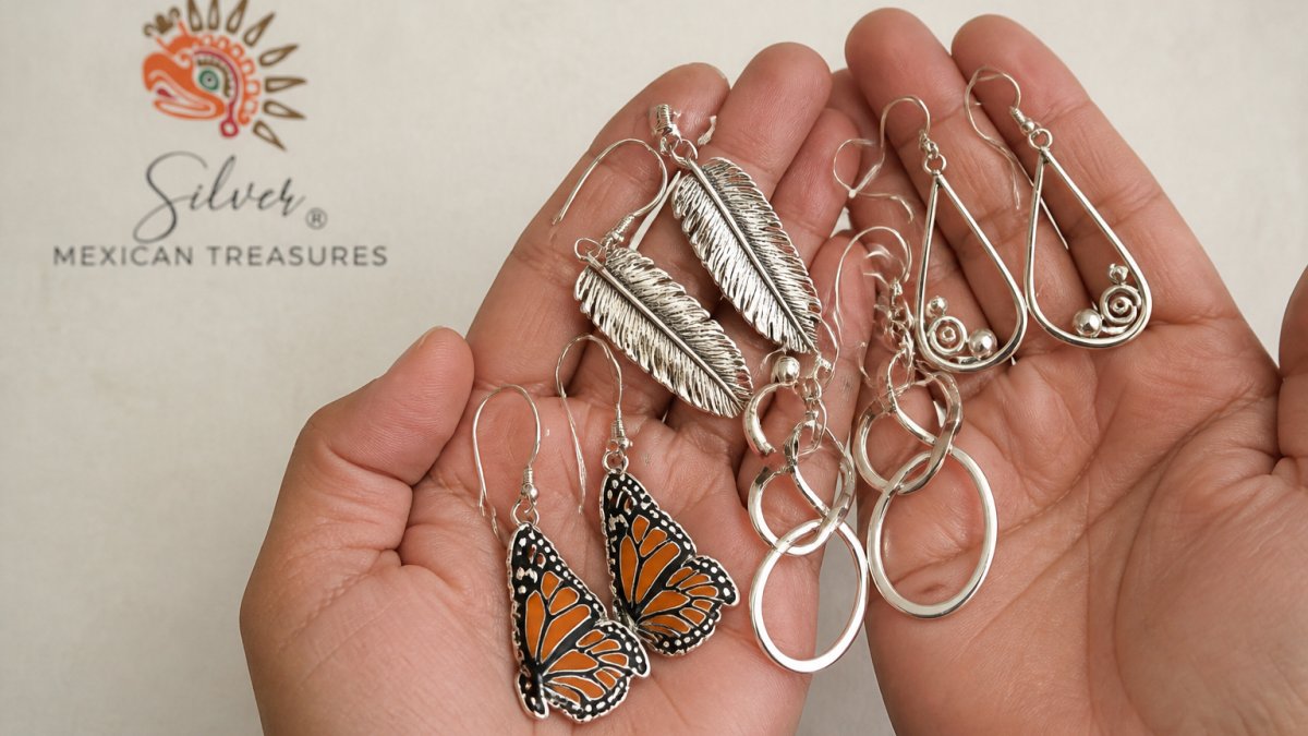 Close-up of hands holding multiple handcrafted Taxco silver earrings with different designs, monarch-inspired drops, feather motifs, sculptural wire forms, showing fine detail and craftsmanship against a neutral cream background