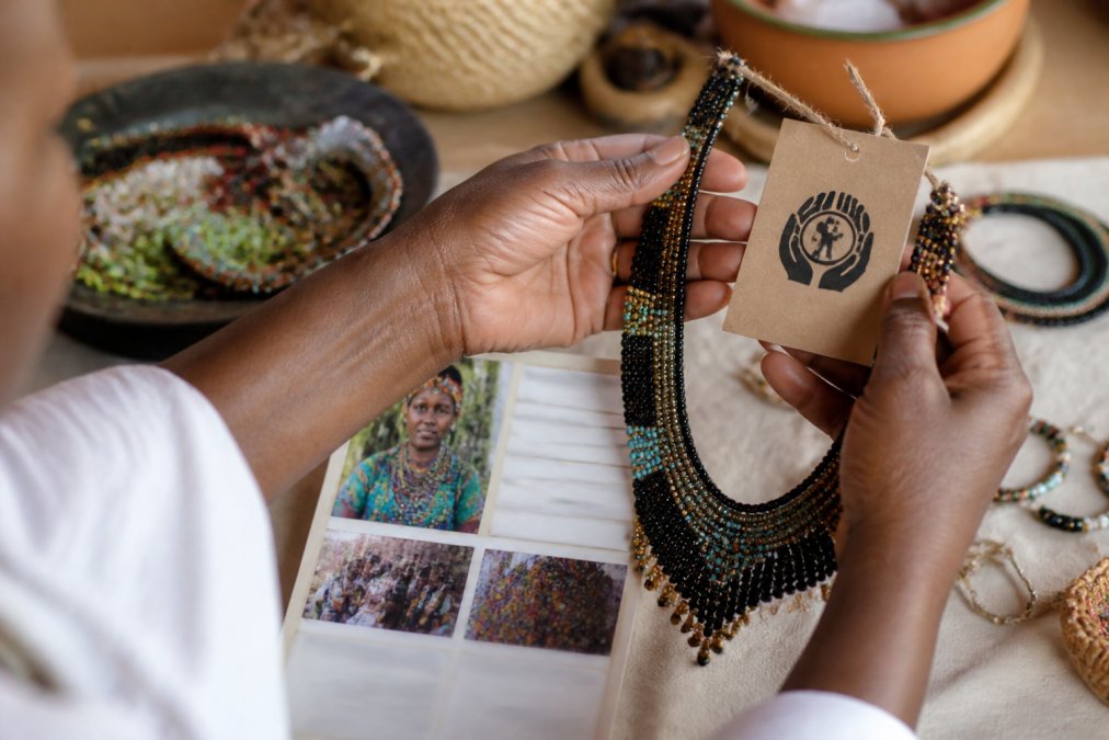 Close-up of hands holding a handcrafted beaded necklace, examining the intricate craftsmanship and beadwork while holding a fair trade certification card and documentation showing artisan name and community details