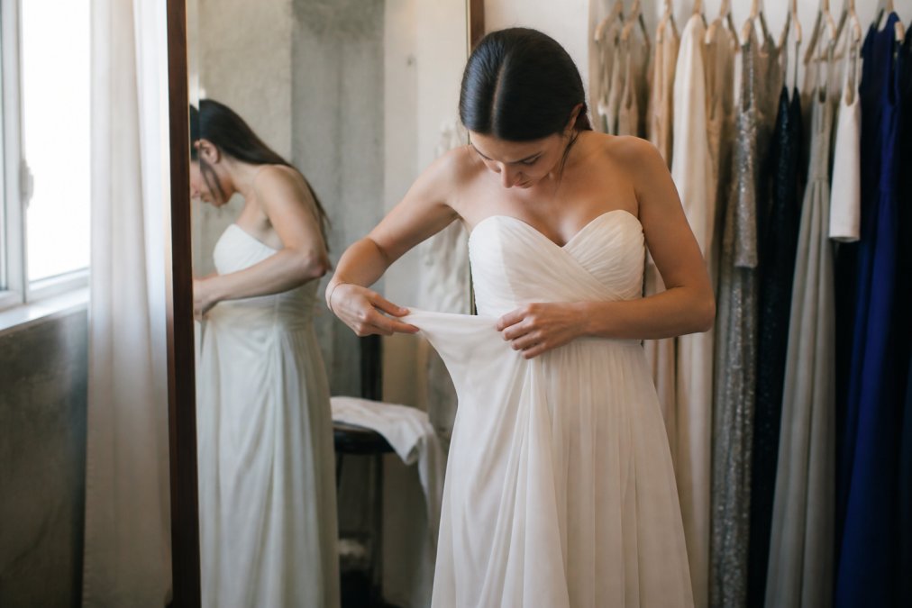 Woman examining a formal dress in detail, checking seams, fabric drape, and construction quality while trying it on in a fitting room with natural lighting