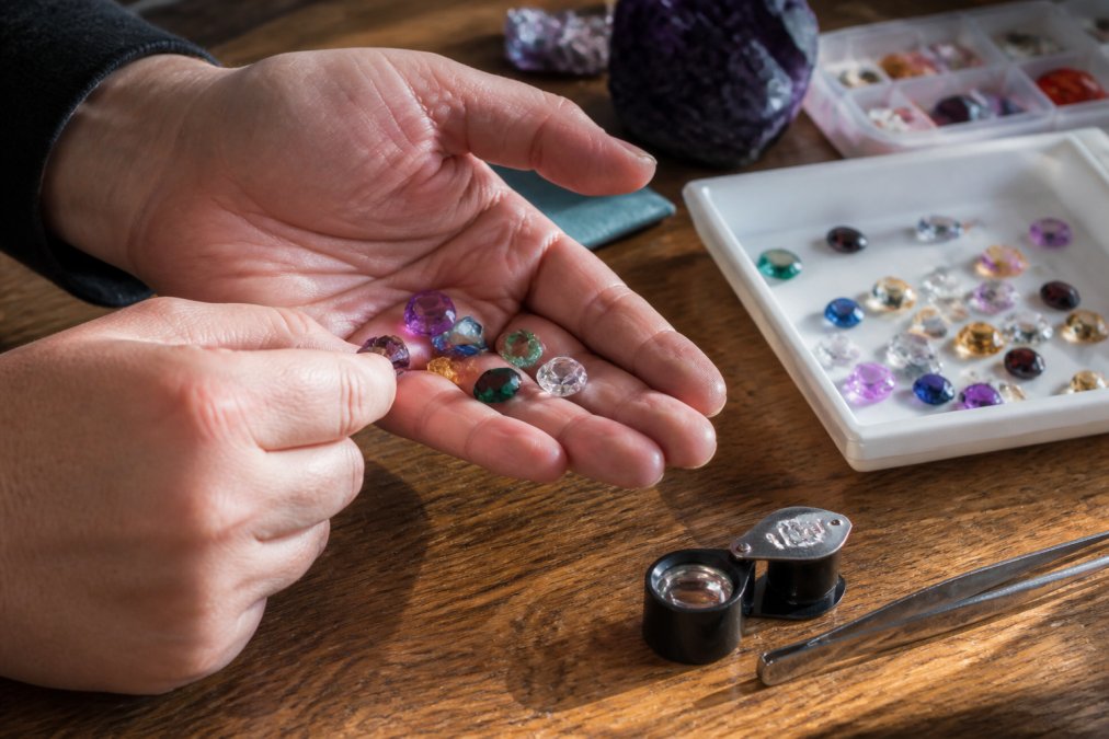 Close-up of a person's hands holding several loose gemstones of different colors up to natural light, examining them carefully on a wooden surface with a loupe magnifier resting nearby