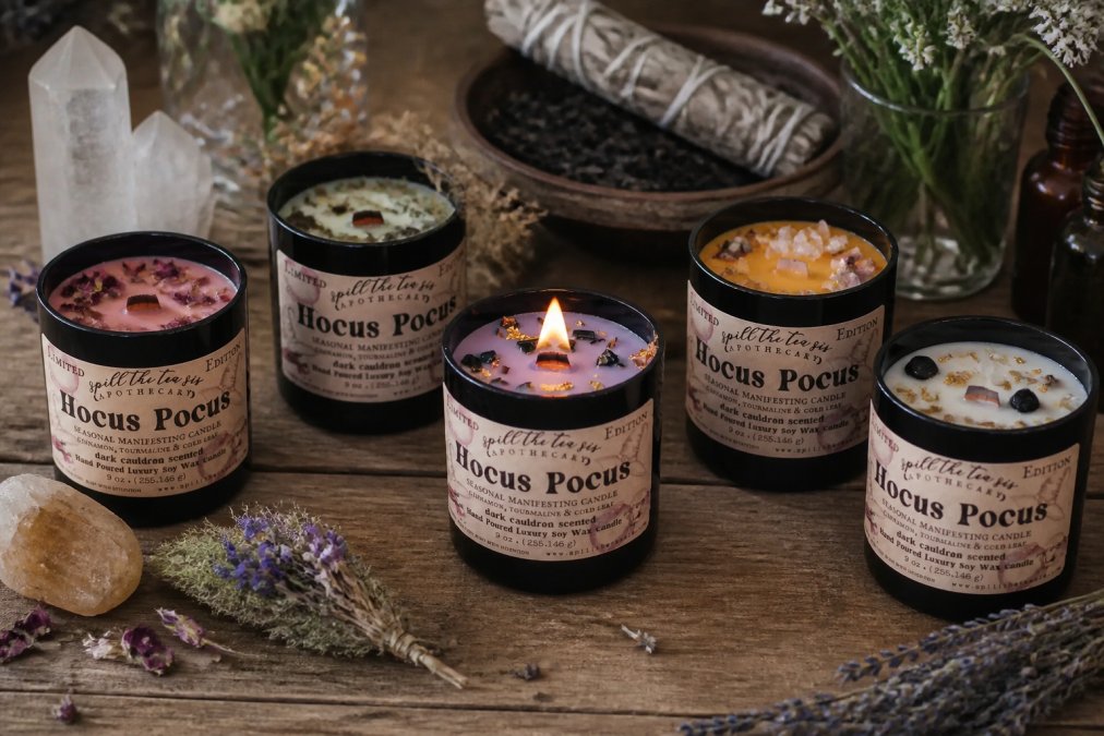 Close-up of several intention candles with different colored wax and labels, arranged on a wooden surface with crystals and dried botanicals nearby, soft natural lighting
