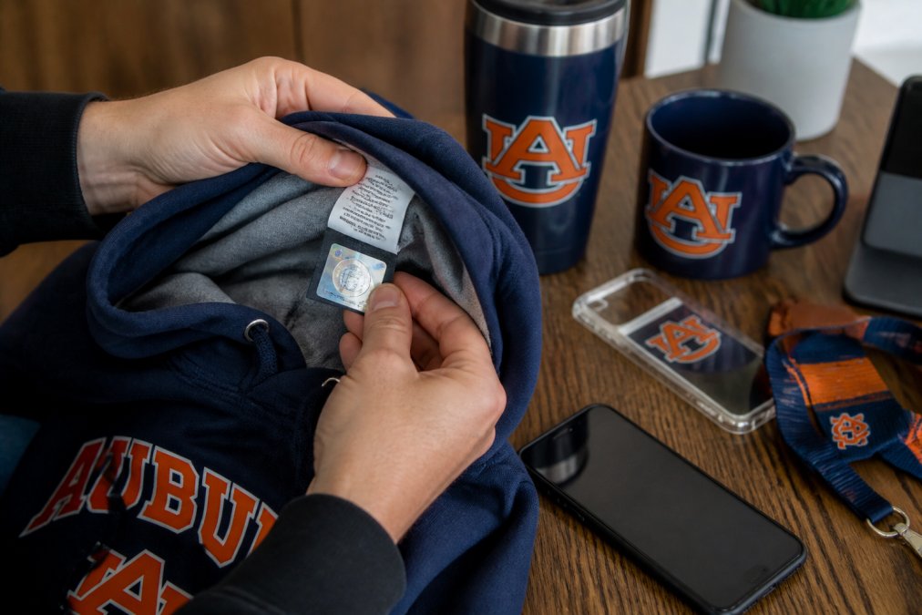 A close-up of a person's hands holding a college hoodie, inspecting the inside hang tag and holographic licensing sticker under bright indoor lighting, with a smartphone showing a team logo verification page nearby