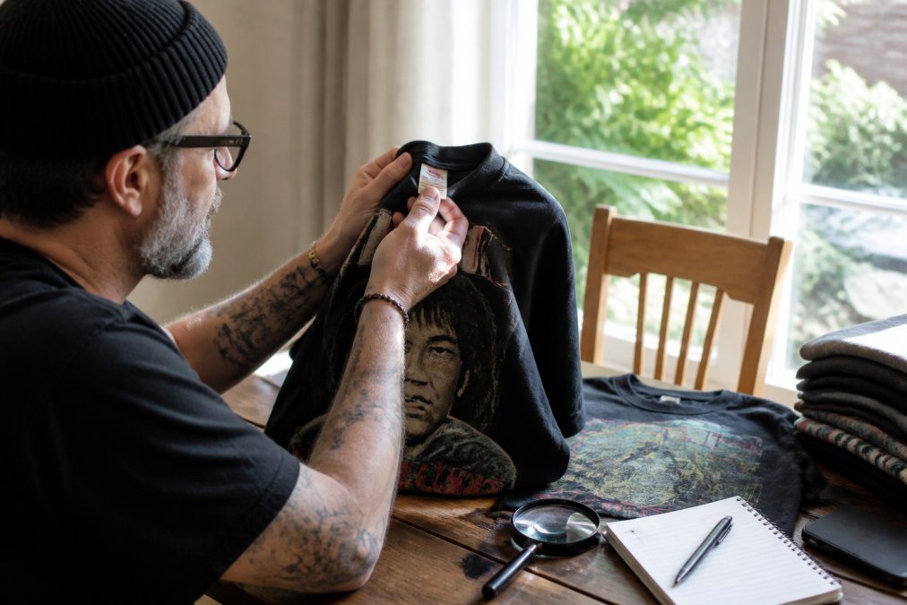 Person carefully inspecting the inside collar tag and screen print detail of a vintage graphic tee, held up to natural window light in a home setting