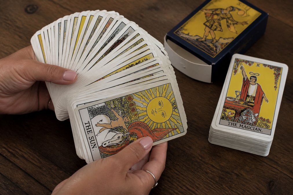 Close-up of hands holding an authentic tarot deck fanned across a wooden table, showing crisp card edges, vibrant artwork detail, and intact box packaging with clear printing