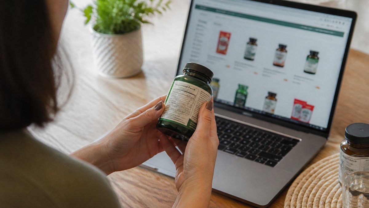 Close-up of a person's hands turning a supplement bottle to read the label, with a laptop showing an online store open on the desk beside it