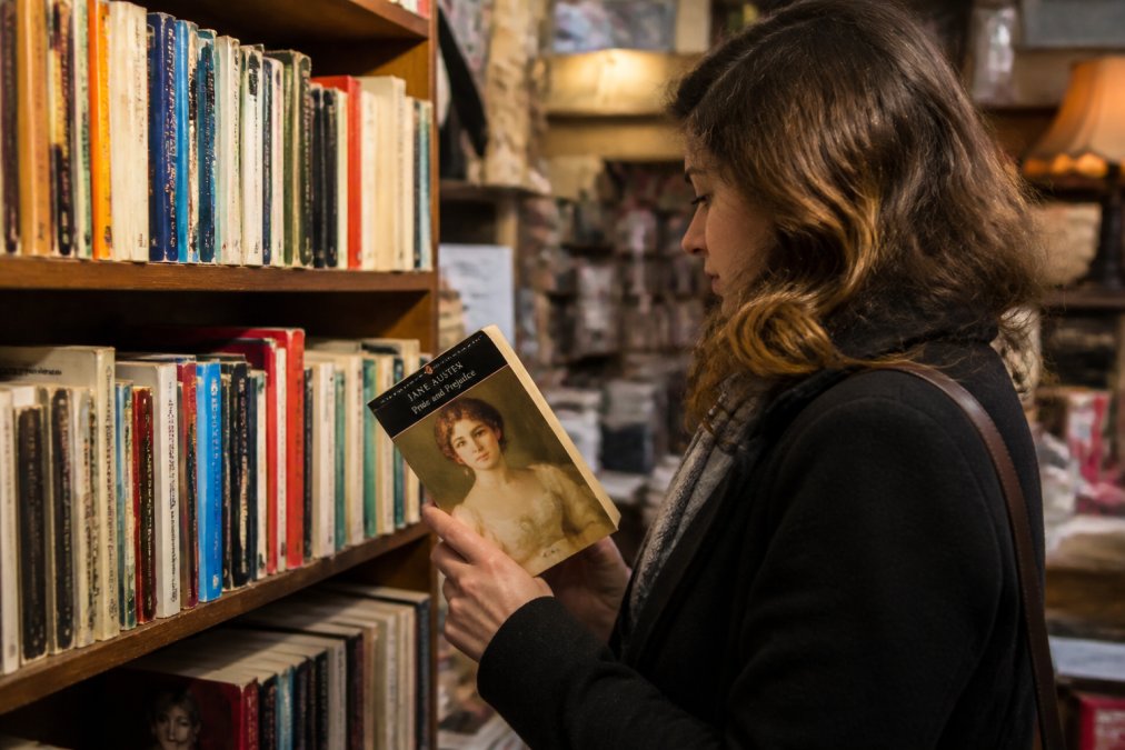 Una persona hojeando una estantería repleta de libros de bolsillo de segunda mano en una tienda benéfica cálida y abarrotada, sacando una novela clásica para examinar la cubierta bajo una suave iluminación cenital