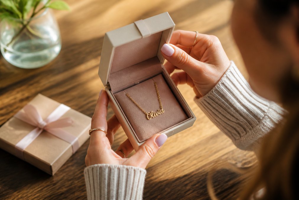 A woman's hands opening a soft-touch jewelry box to reveal a personalized name necklace, warm natural light on a wooden table