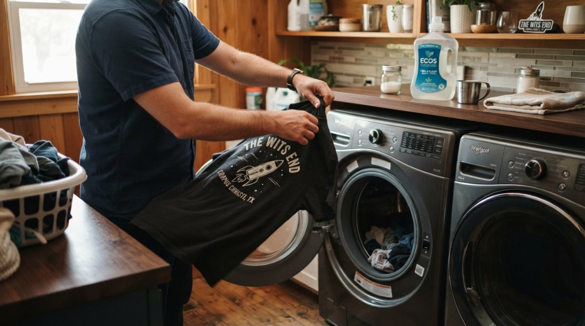 Person turning a dark graphic tee inside out before placing it into a front-loading washing machine, with a bottle of gentle, fragrance-free detergent visible on the laundry shelf in the background, warm indoor lighting