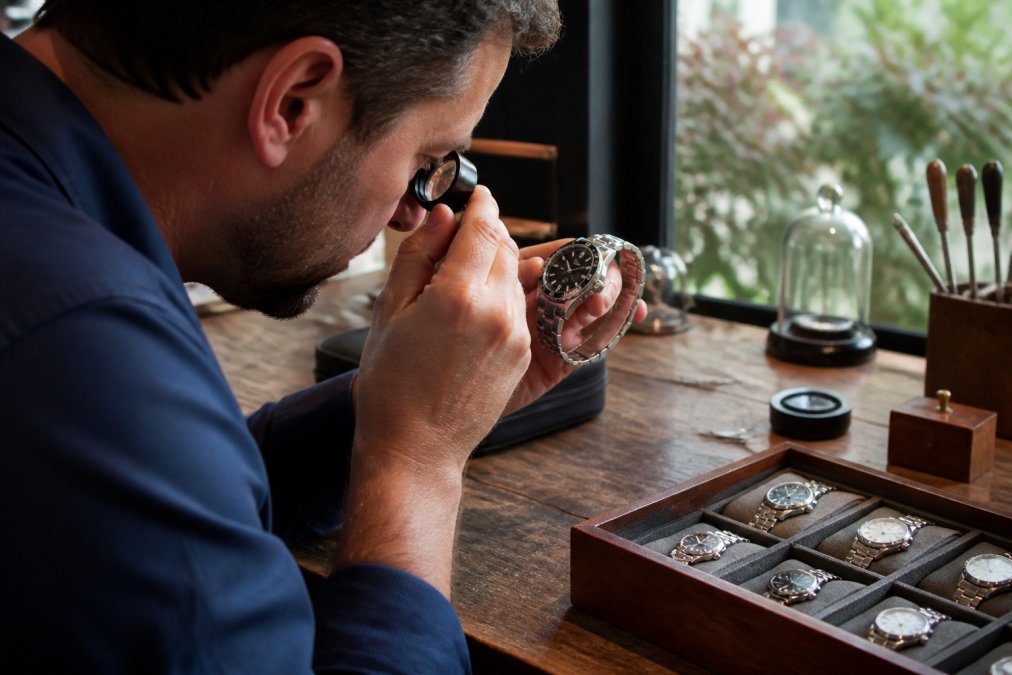 Person examining a pre-owned luxury watch with magnifying glass, checking condition and authenticity details on a wooden workbench with natural daylight