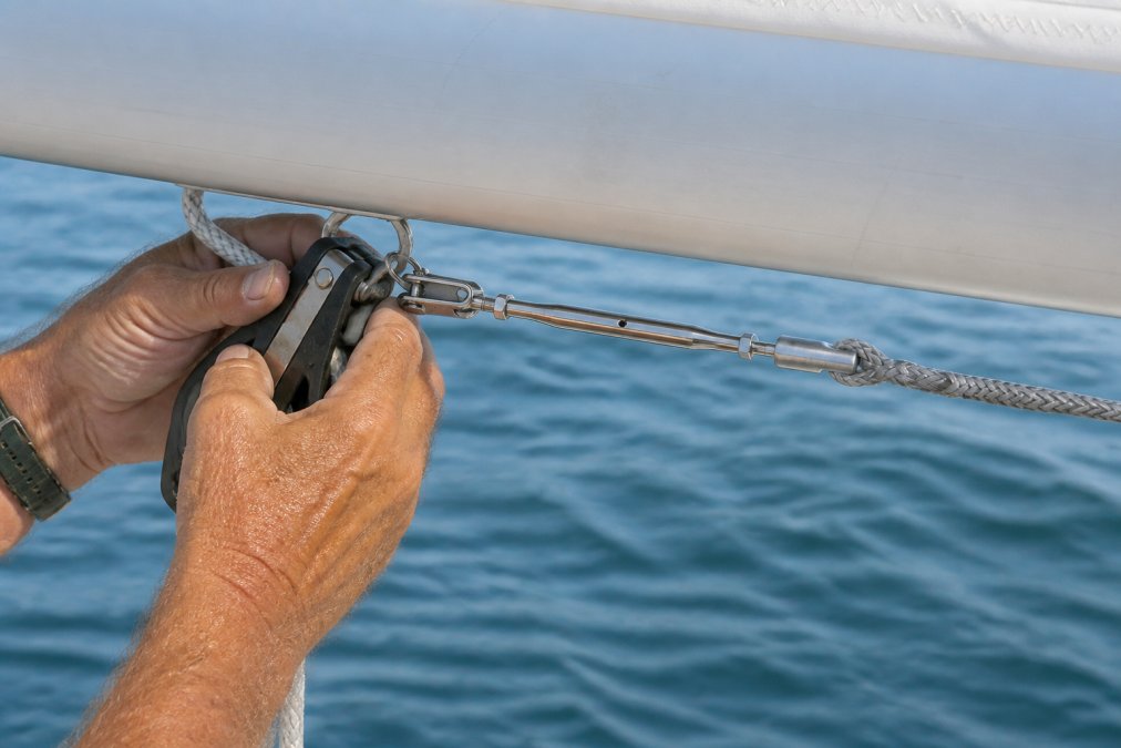 Sailor's weathered hands manipulating sheaves and blocks on sailboat boom during rigging deployment, with furling line visible and blue water in background