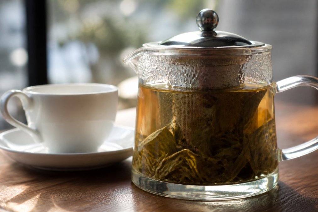 Close-up of loose leaf tea steeping in a glass infuser with hot water, showing full leaves expanding, with a white ceramic cup and saucer beside it on a wooden table with soft natural morning light