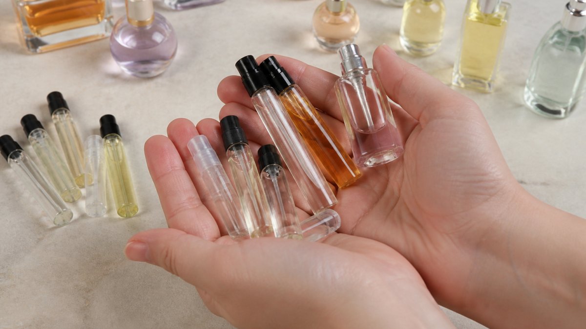 Close-up of hands holding multiple perfume sample vials and miniature bottles in various sizes and colors arranged on a neutral surface, showing the variety available